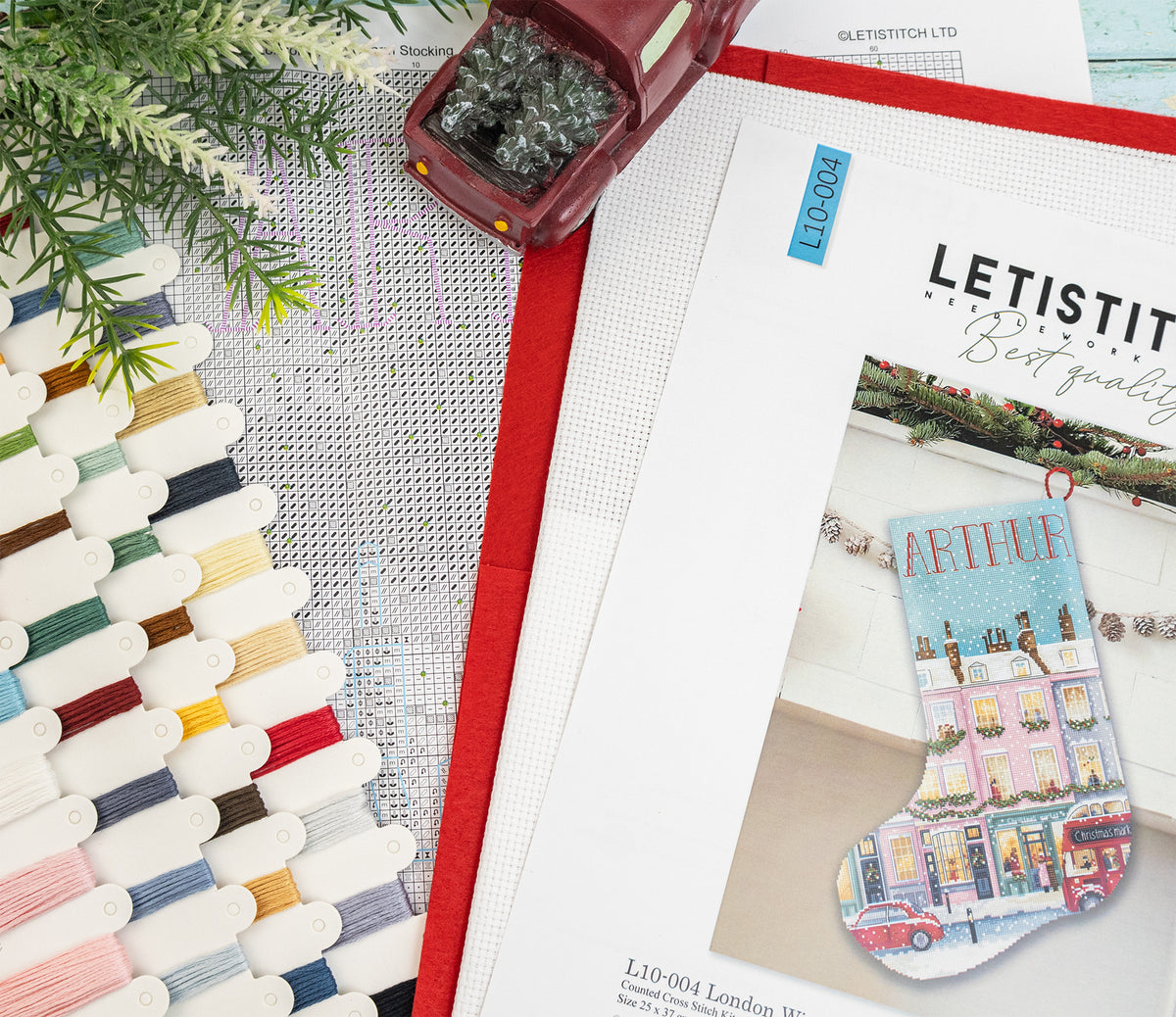 A flat-lay of a cross-stitch kit showing a printed pattern booklet with a colorful Christmas stocking design, alongside spools of thread, a red fabric trim, and a small decorative tree on a white mesh background.