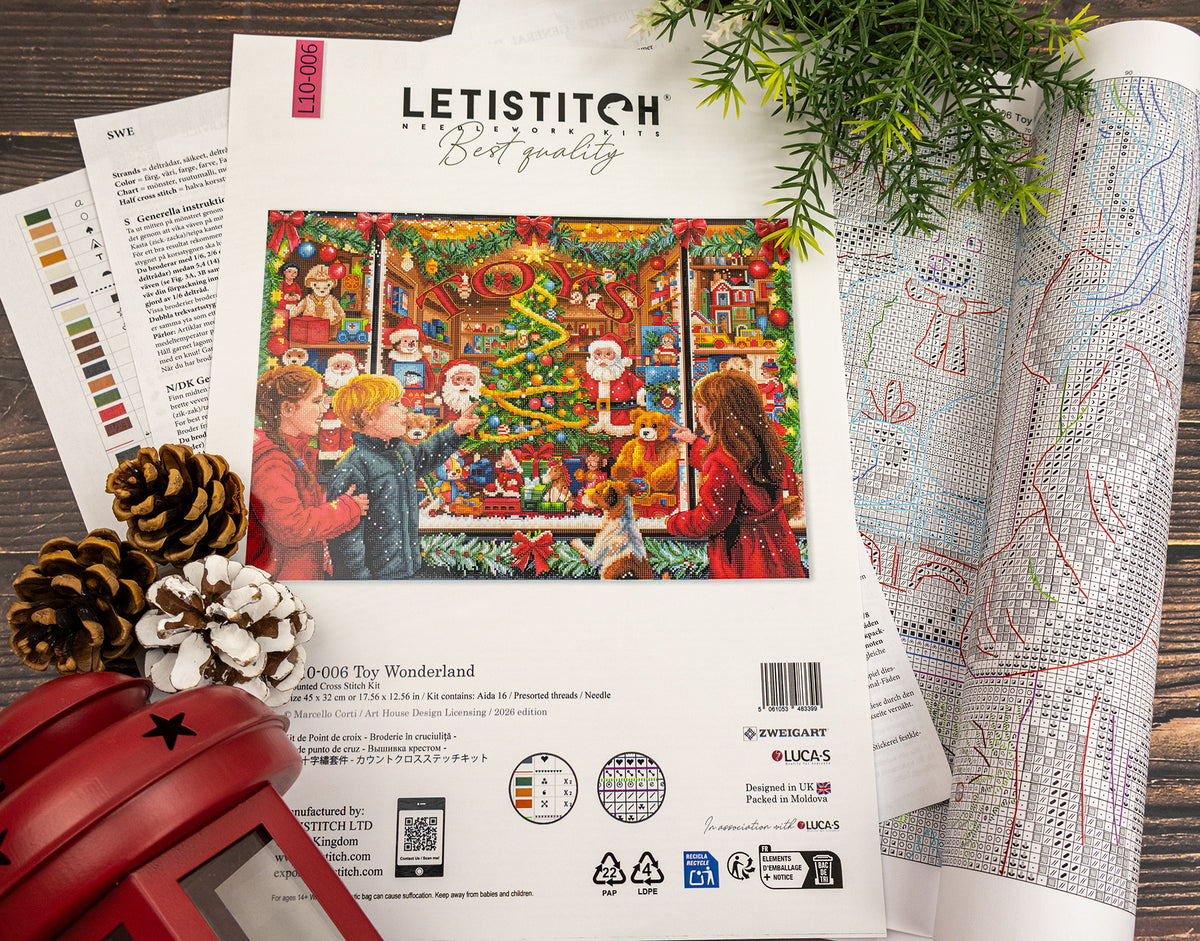 A flat-lay of a Letistitch cross-stitch kit cover for 'Toy Wonderland', surrounded by open pattern sheets, pinecones, and evergreen branches on a dark wooden surface.