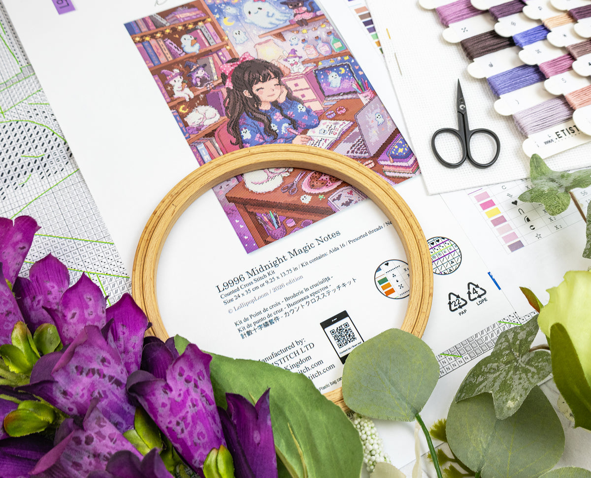 A flat-lay of craft supplies including a wooden embroidery hoop, printed pattern sheets, purple flowers, green leaves, thread spools, and black-handled scissors on a white background.