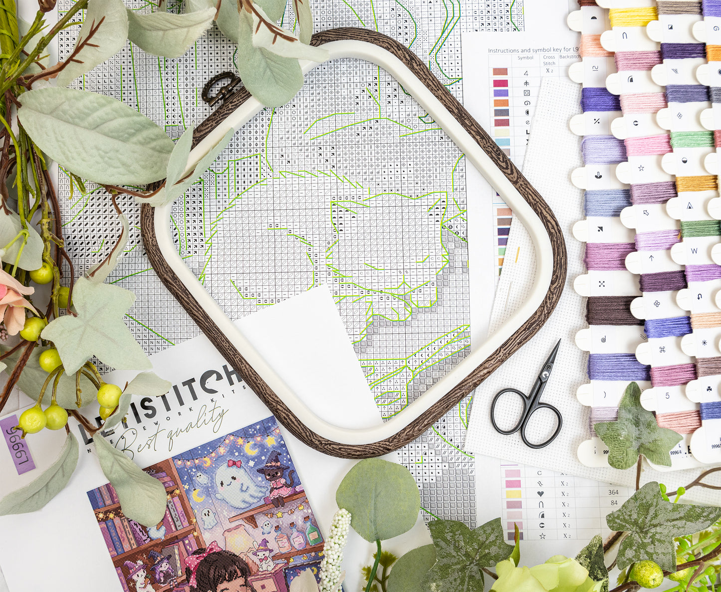 A styled flat-lay of embroidery supplies, featuring a white hoop with a brown wooden frame, a flocked cat embroidery pattern, a color swatch chart, scissors, and green foliage.