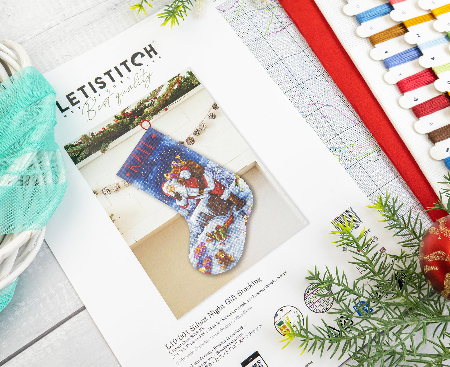 A printed sewing pattern booklet for a Letistitch Christmas stocking lies on a light wood surface, surrounded by craft props including spools of thread, a red ribbon, green pine branches, and a small ornament. The cover shows a completed blue stocking with festive embroidery.
