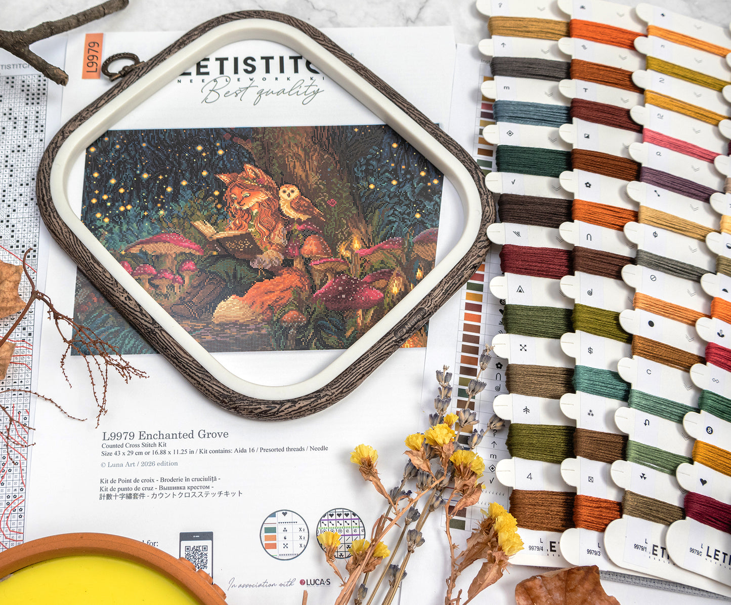 Embroidery hoop with a colorful design on fabric, surrounded by spools of thread and dried flowers.