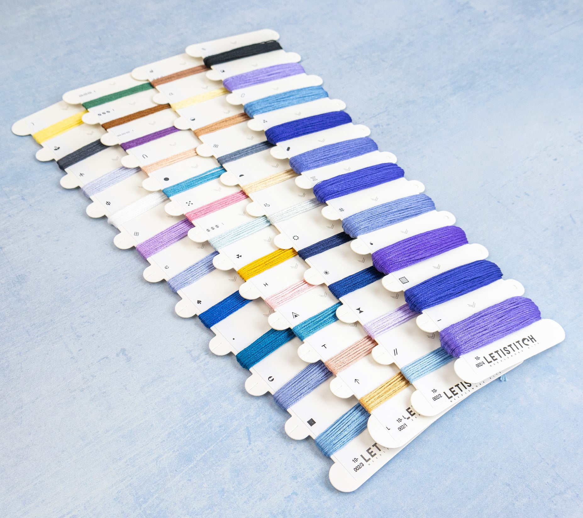 A set of embroidery floss thread spools arranged in rows on a light blue textured background. The threads include a wide range of colors such as blues, purples, pinks, yellows, greens, and browns, each mounted on a white card labeled 'LETISTITCH'.