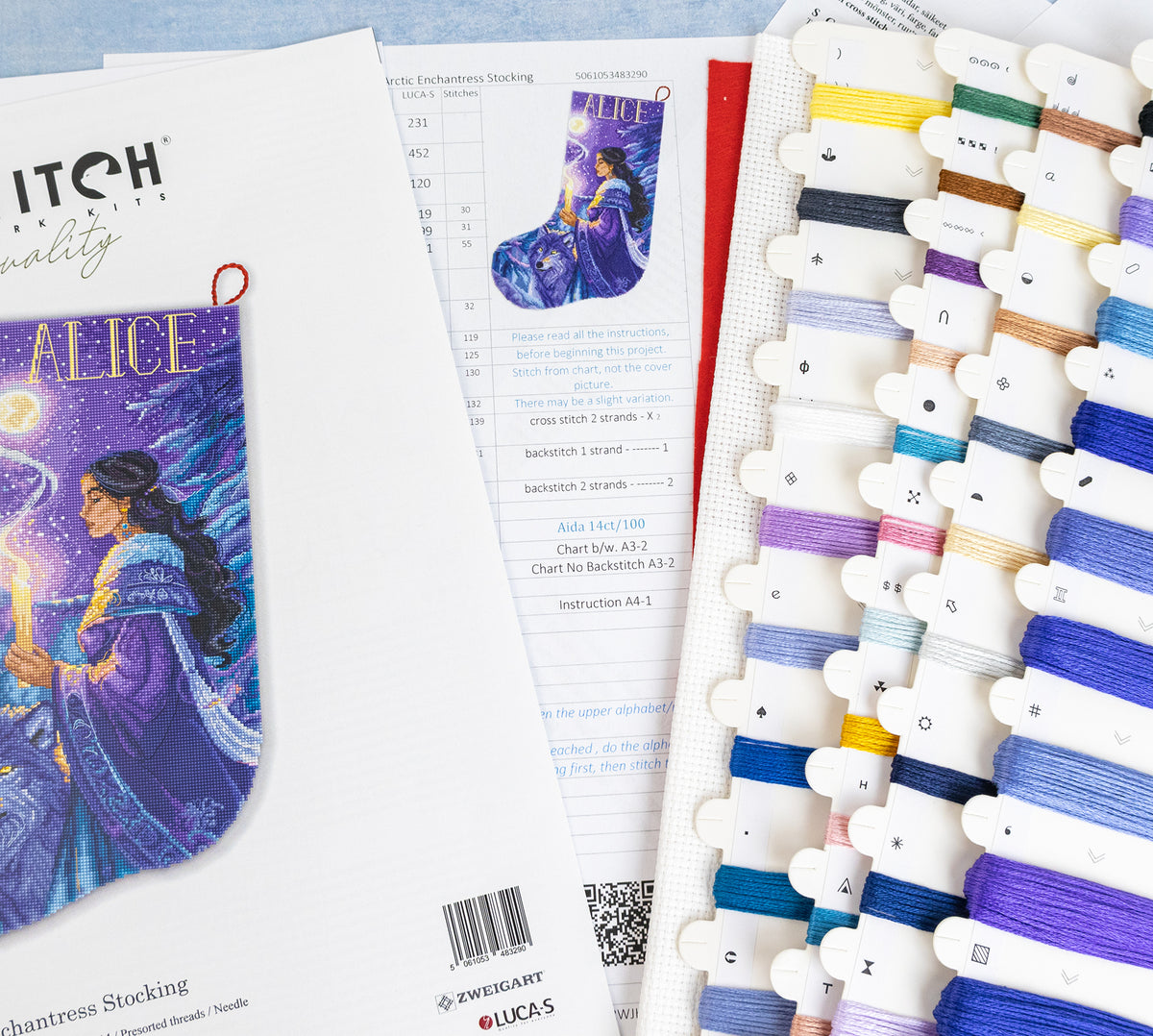 A flat lay of craft supplies for Enchanted Stocking kit, showing the printed pattern booklet, a finished stocking with a purple illustration, an instruction sheet, and multiple spools of thread in various colors.