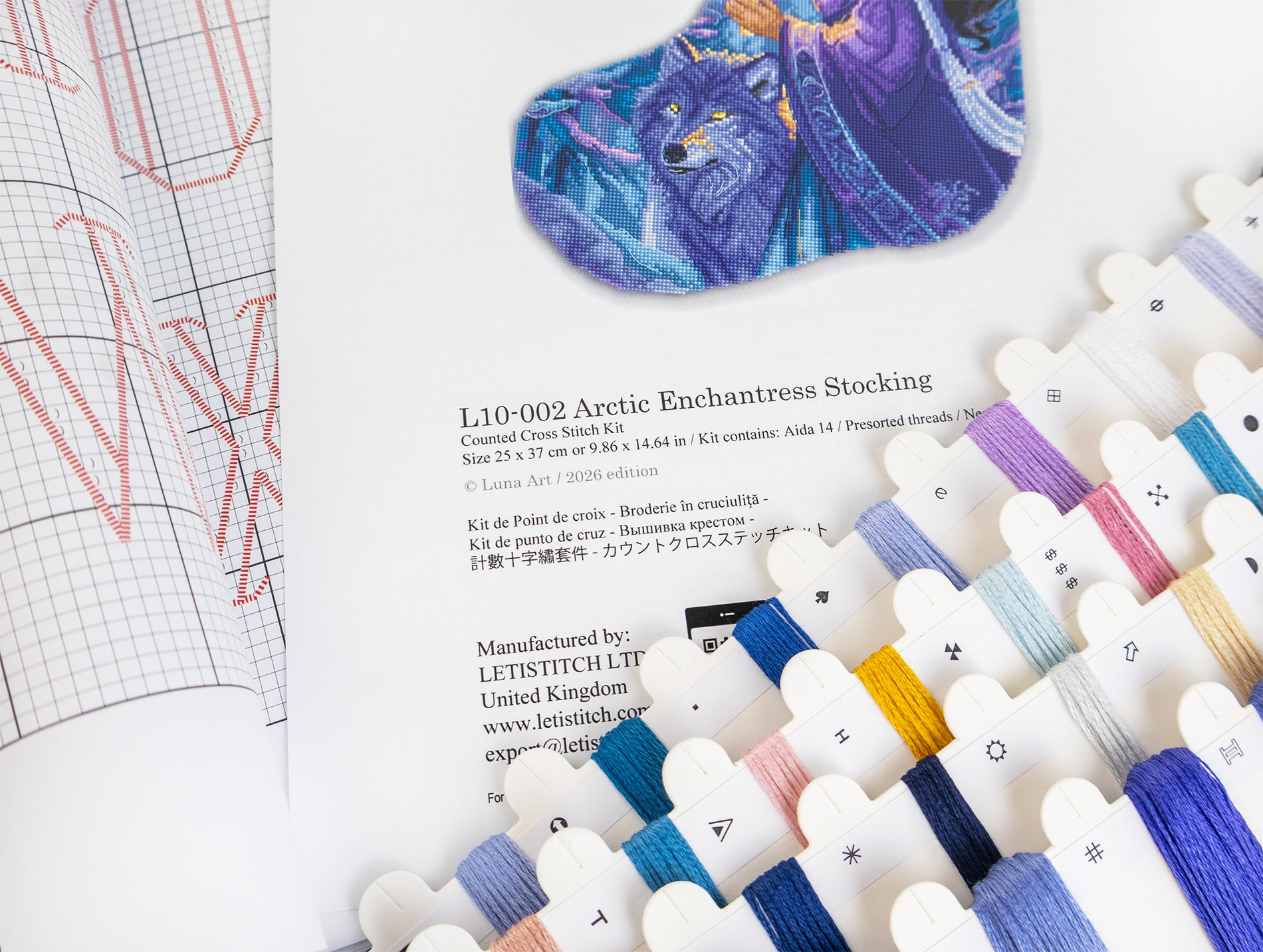 An embroidery kit booklet page showing the pattern design of an Arctic Enchanted Stocking, alongside a colorful dragon illustration and a row of white embroidery floss skeins in various colors.
