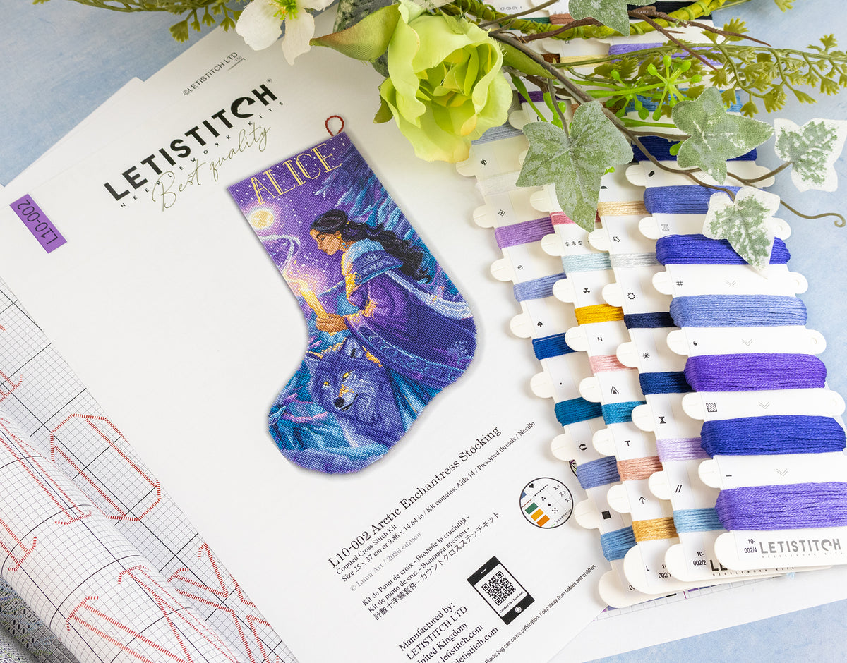 A flat-lay of a Letistitch embroidery kit showing the printed design sheet for an Alice stocking, a swatch card of purple and blue embroidery floss, and decorative flowers on a light blue surface.