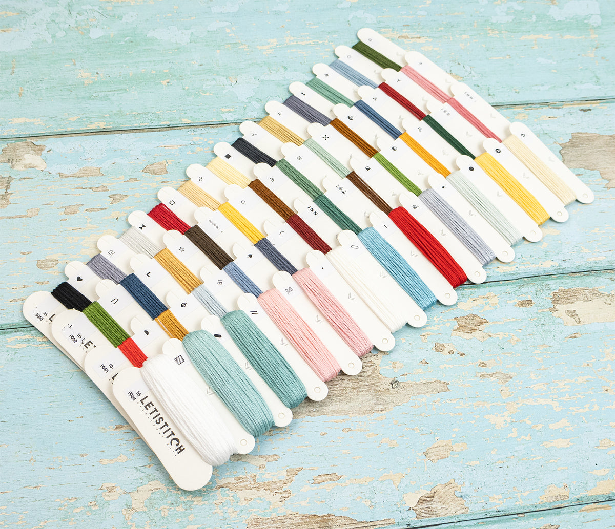 A set of embroidery floss spools arranged in rows on a distressed light blue wooden surface. The floss comes in many colors, including black, green, red, yellow, blue, pink, brown, and white, with the brand name 'LETISTITCH' visible on several spools.