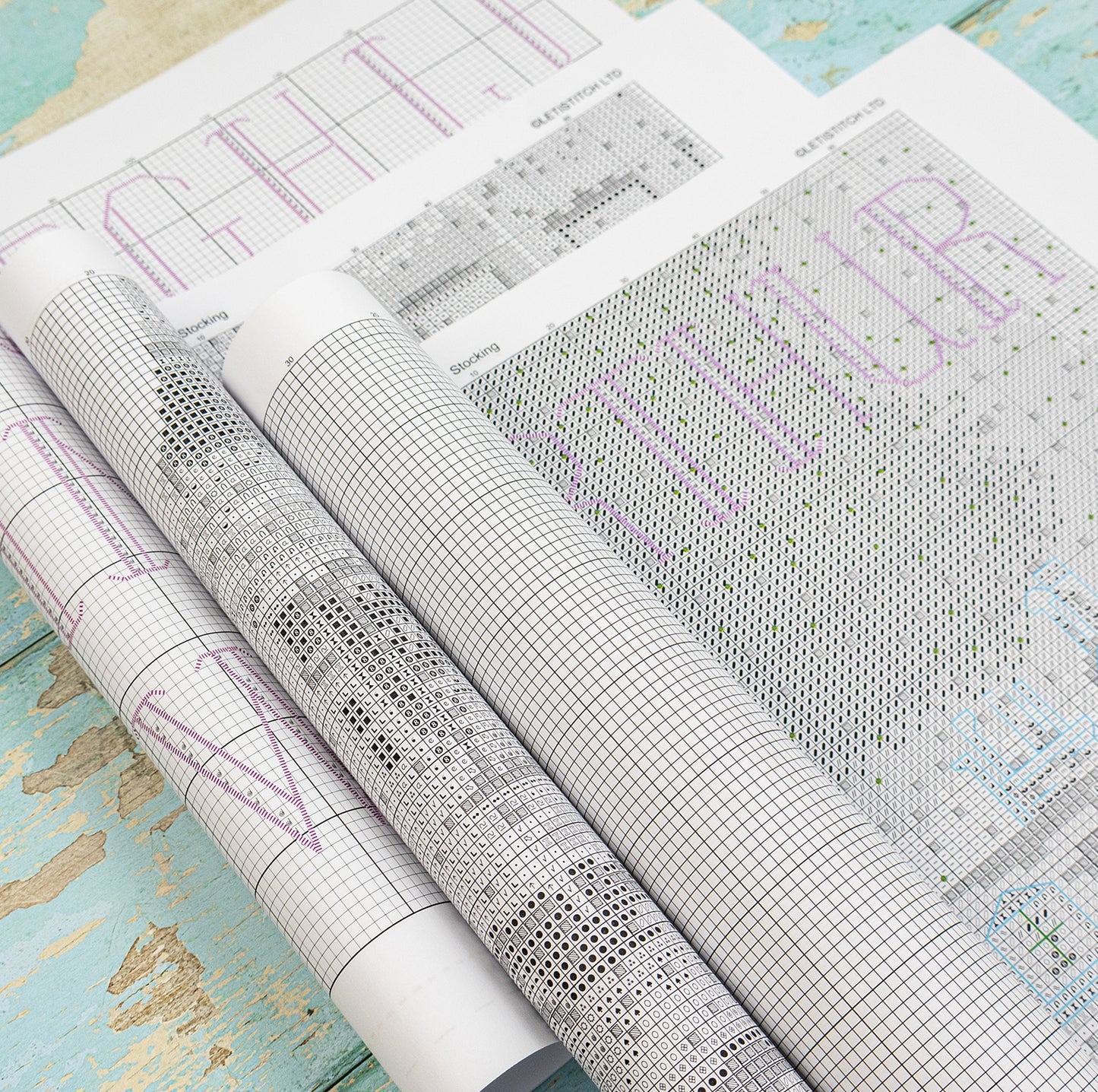 Rolled and unrolled patterned quilting charts on a distressed turquoise wooden surface. The charts feature black grid lines, black and gray floral and geometric motifs, and purple text