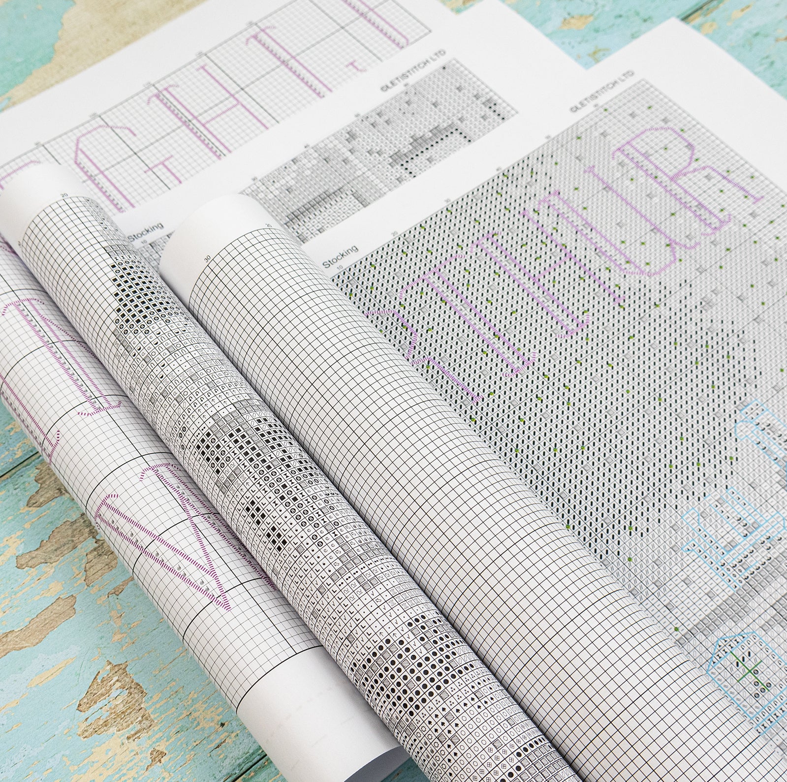 Rolled and unrolled patterned quilting charts on a distressed turquoise wooden surface. The charts feature black grid lines, black and gray floral and geometric motifs, and purple text