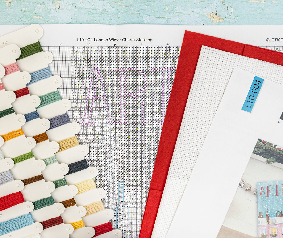 A close-up flat lay of needlepoint or cross-stitch materials, including a printed design sheet with the word ART, a red border sheet, white mesh grids, and multiple thread spools in assorted colors arranged along the left side.