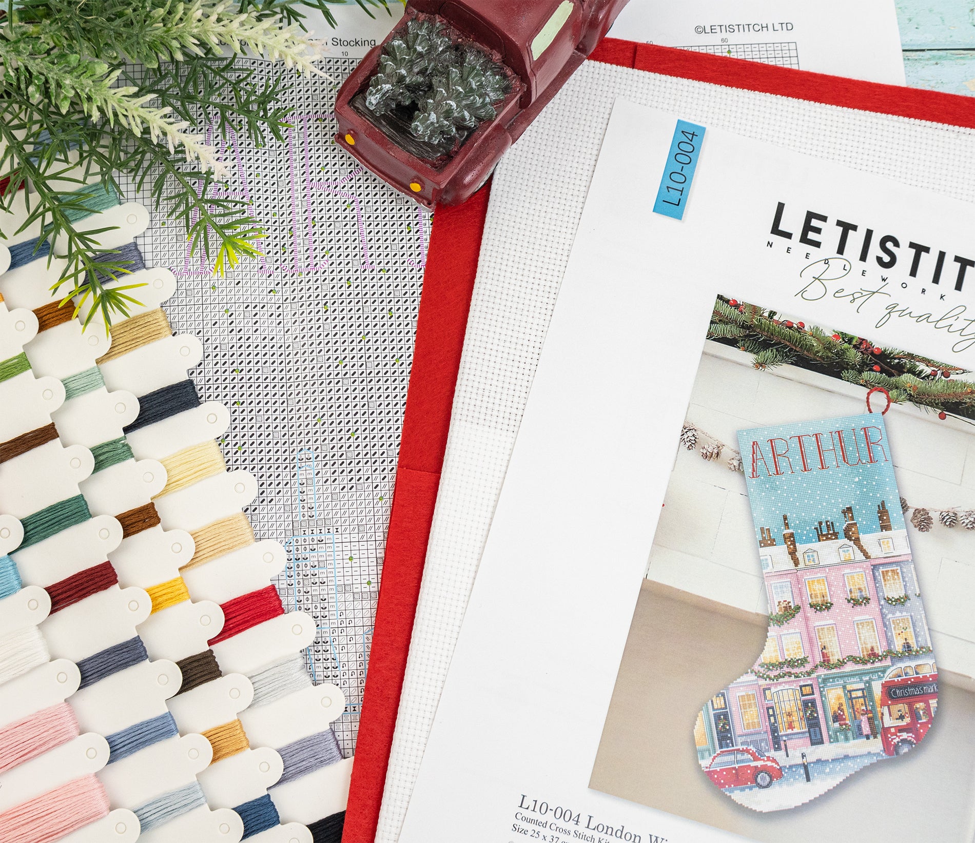 A flat-lay of a cross-stitch kit showing a printed pattern booklet with a colorful Christmas stocking design, alongside spools of thread, a red fabric trim, and a small decorative tree on a white mesh background.