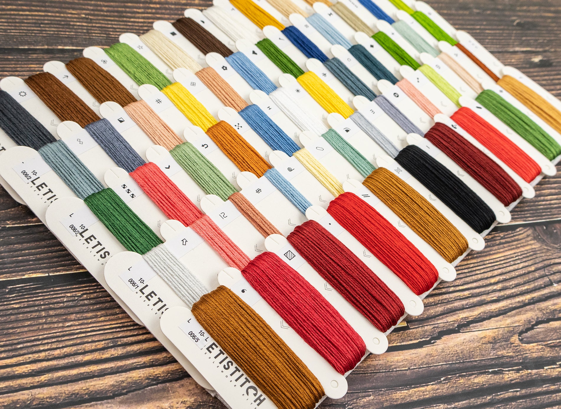 A set of Letistitch embroidery floss thread spools arranged in neat rows on a dark wood background, showcasing a wide variety of colors including red, green, blue, yellow, brown, and black.