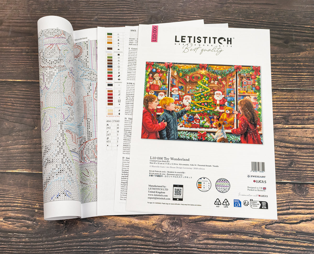 An open Letistitch needlework kit booklet showing the front cover with a colorful Christmas scene and an open pattern page with color charts, placed on a dark wood surface.