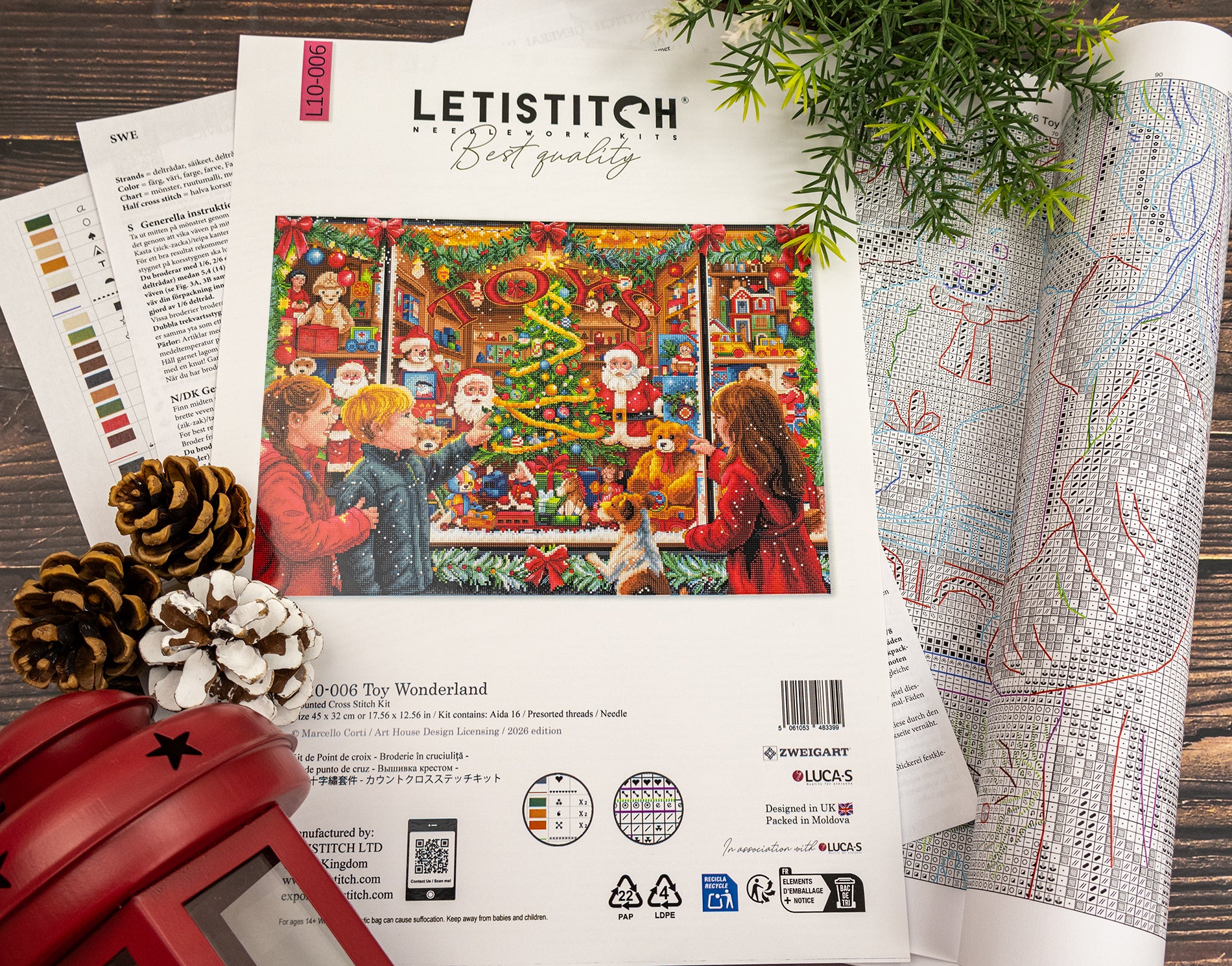 A flat-lay of a Letistitch cross-stitch kit cover for 'Toy Wonderland', surrounded by open pattern sheets, pinecones, and evergreen branches on a dark wooden surface.