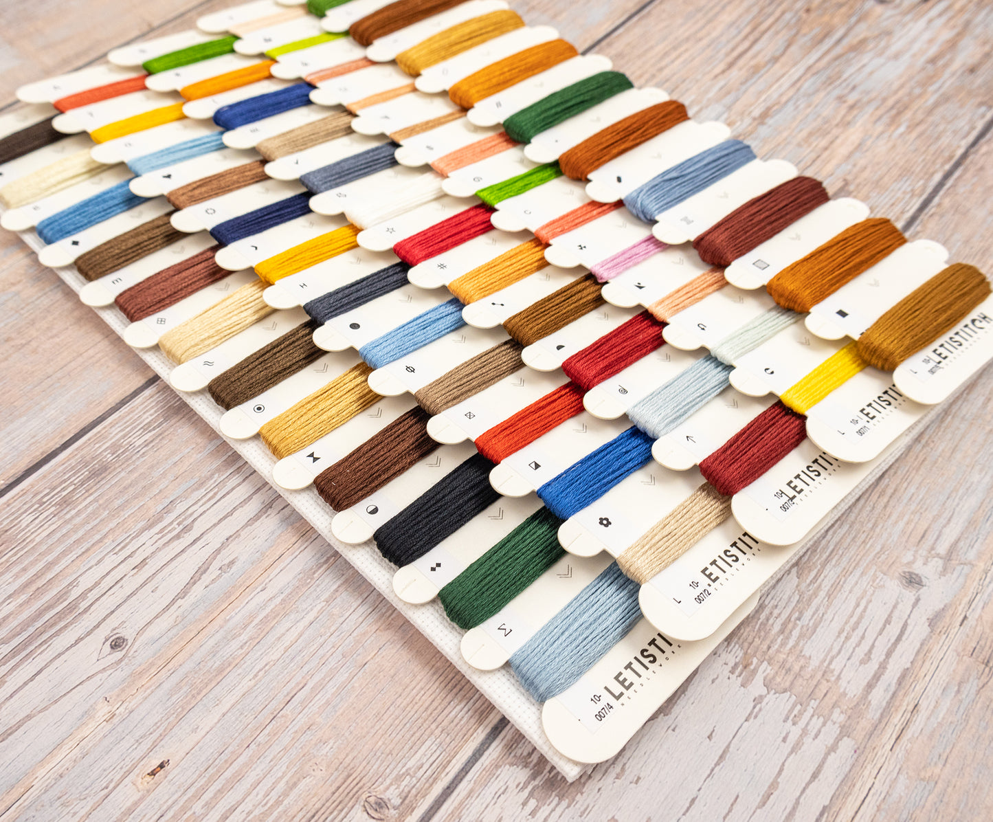 A wooden embroidery floss display board filled with many thread spools in assorted colors, photographed on a light wood tabletop. The brand name LETISTITCH is visible on the spool tabs.