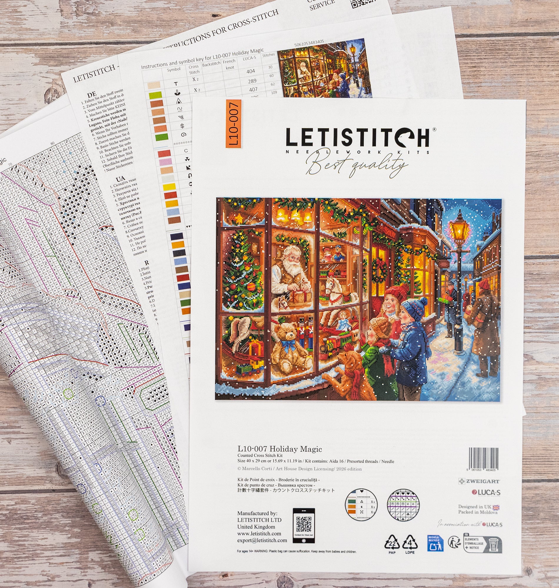 A Letistitch cross-stitch kit package labeled 'Holiday Magic' lies on a light wood surface, partially covering a folded fabric sample and a color chart. The package features a festive illustrated scene of a snowy street with people and a decorated window.