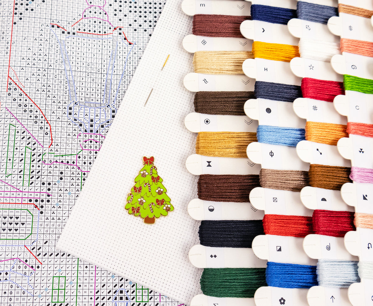 A close-up flat lay of embroidery supplies: a partially completed cross-stitch pattern sheet, a white fabric panel with a small embroidered green Christmas tree, and multiple spools of thread in assorted colors arranged along the right side.