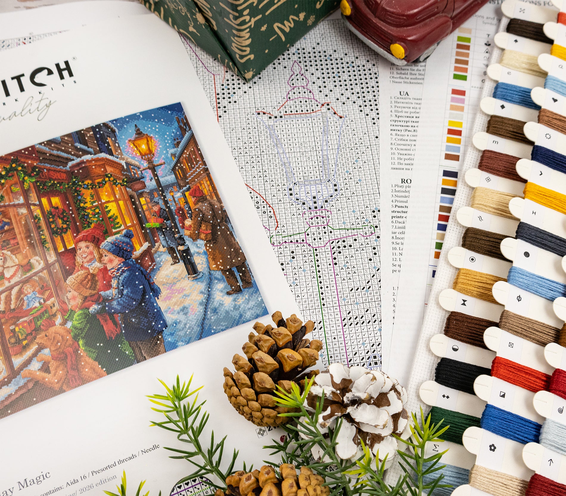 A styled flat-lay of needlework supplies and a finished craft design. On the left is an open book or booklet showing a colorful winter street scene