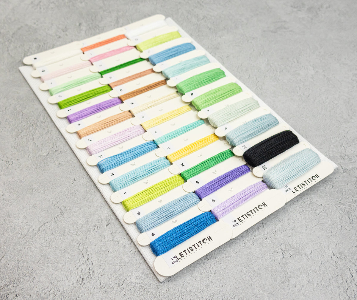 A set of embroidery floss thread spools arranged on a white display card, photographed on a light gray textured surface. The threads include many pastel and bright colors, with one black spool and several pale blue shades.