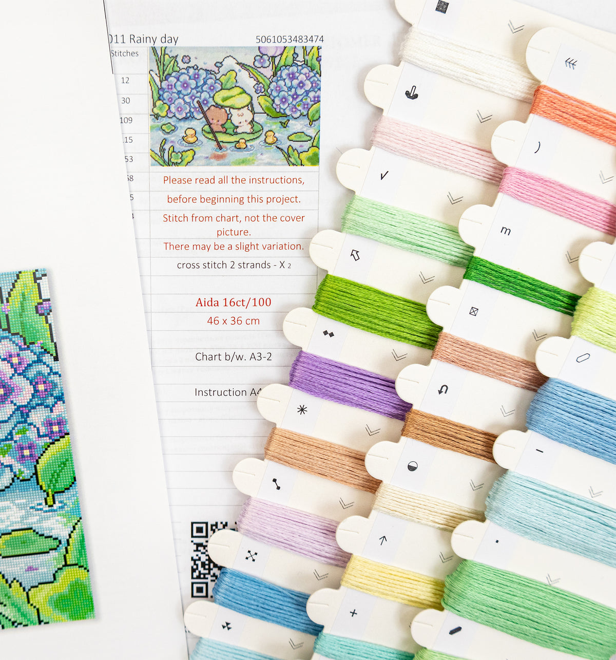 A flat-lay view of a cross-stitch kit showing a printed pattern sheet with instructions and a finished colorful embroidered patch, alongside multiple wooden spools of thread in pastel and bright colors.