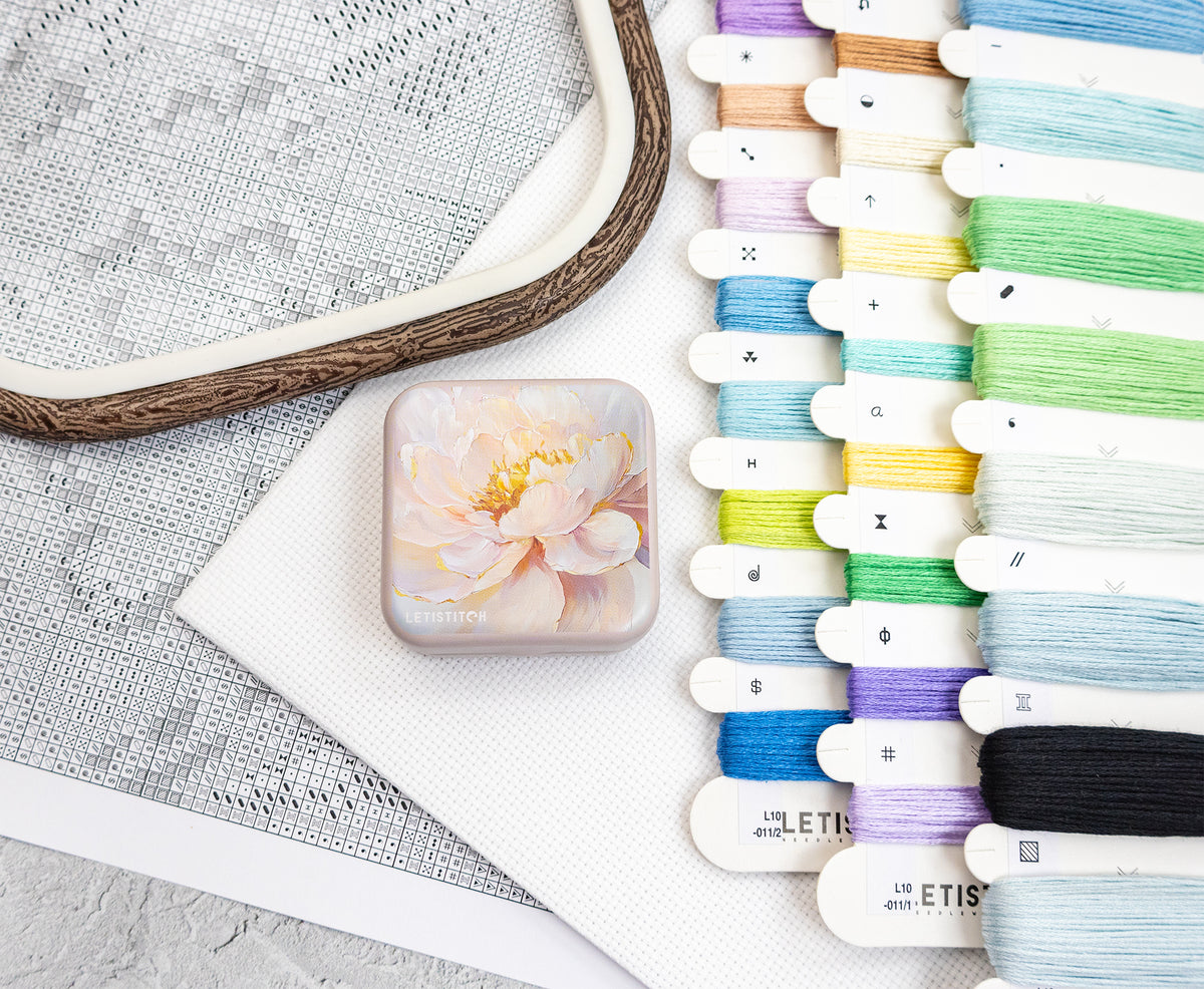 A square aluminum tin box featuring a printed peony flower design, styled on a white textured fabric next to a wooden embroidery hoop and a vertical row of colorful embroidery floss spools.