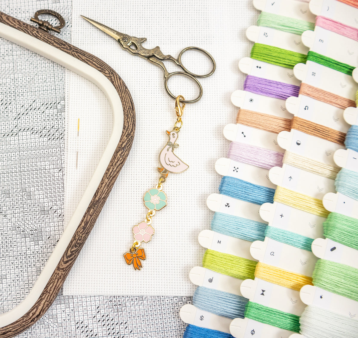 A styled flat-lay of embroidery supplies on a white mesh fabric background, including a wooden embroidery hoop, silver embroidery scissors, a decorative enamel bird charm, and multiple spools of thread in pastel colors.