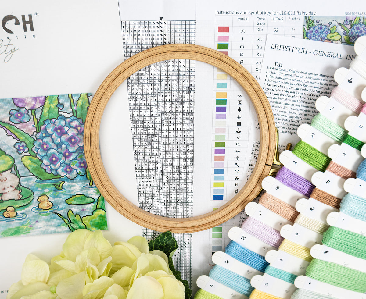 A wooden embroidery hoop holding a cross-stitch pattern chart, surrounded by colorful thread spools, a floral print sheet, and a yellow flower on a white background.