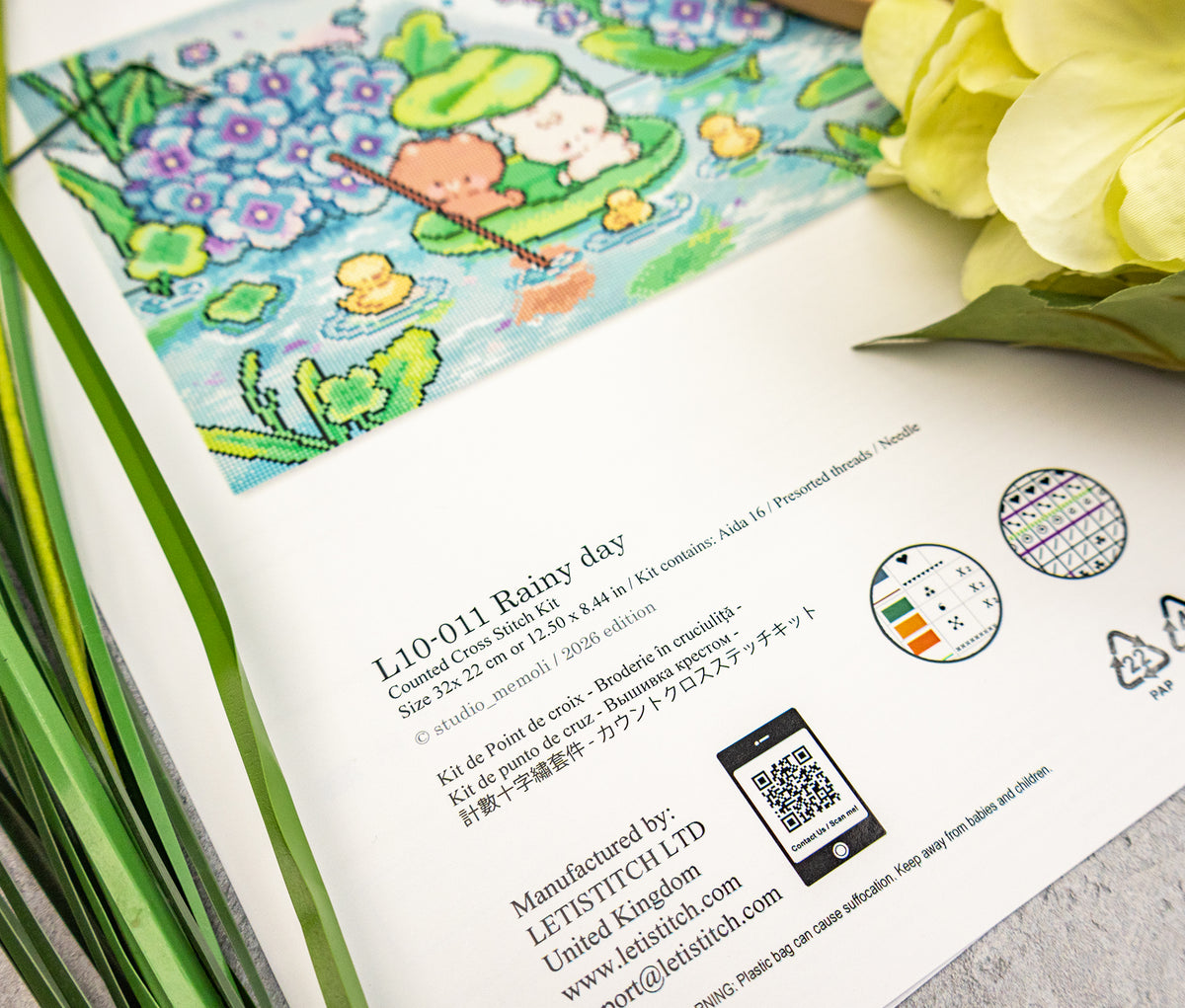 A close-up of a printed kit insert for a craft embroidery design titled 'Rainy day' by LETISTITCH LTD. The insert features a colorful illustration of a frog sitting on a lily pad in a pond, along with product details, a QR code, and material information.