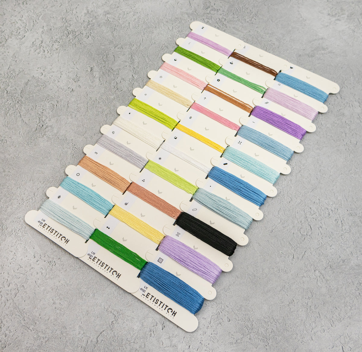 A set of embroidery floss spools arranged in rows on a light gray textured background. The floss comes in many colors including green, pink, yellow, blue, purple, brown, orange, and black, each on a white card labeled 'LETISTITCH'.