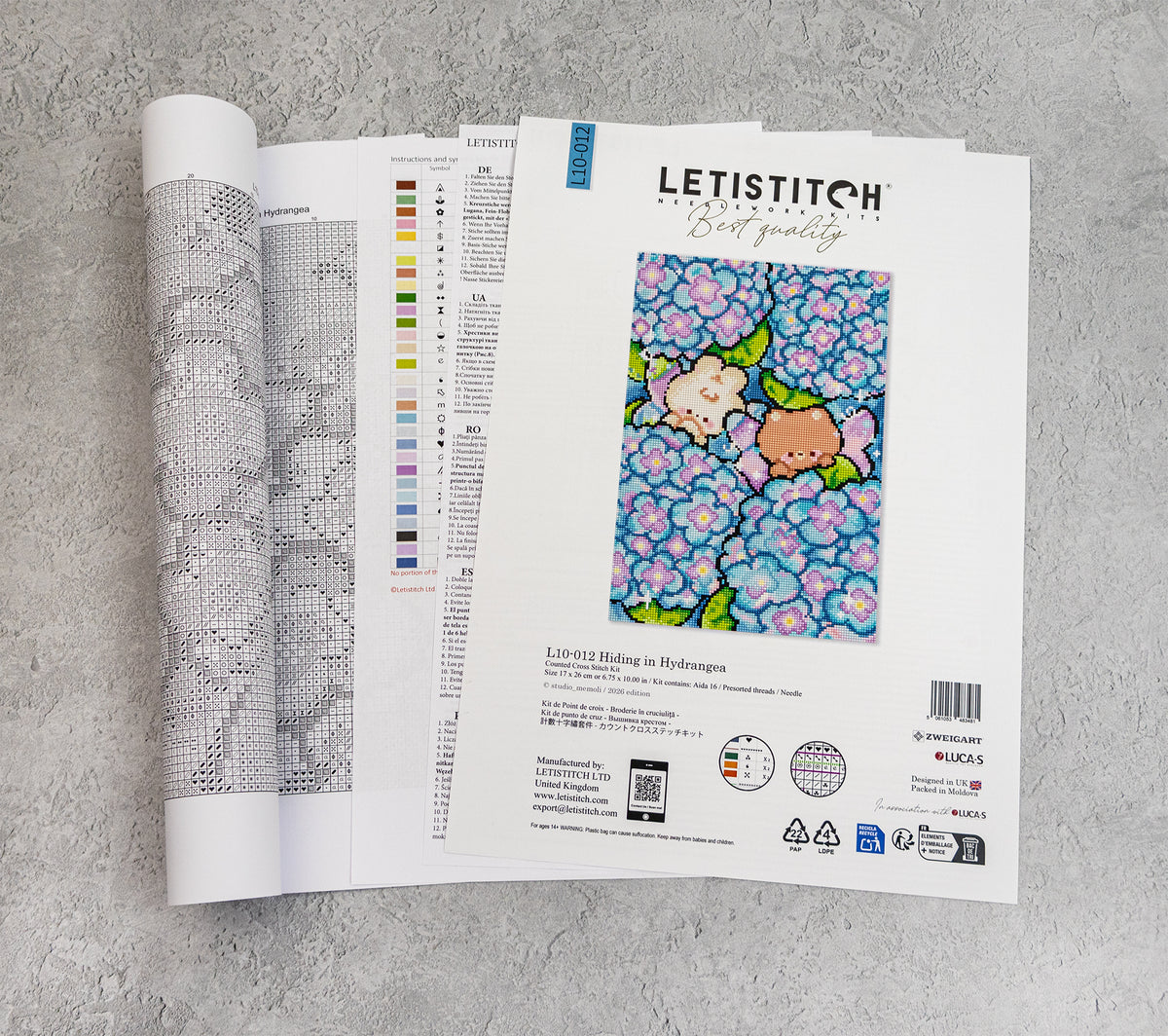 An open Letistitch embroidery pattern booklet lies on a light gray textured surface. The right page shows the cover with the Letistitch logo, a colorful floral sample design, and product details; the left page is partially open, showing the chart layout.