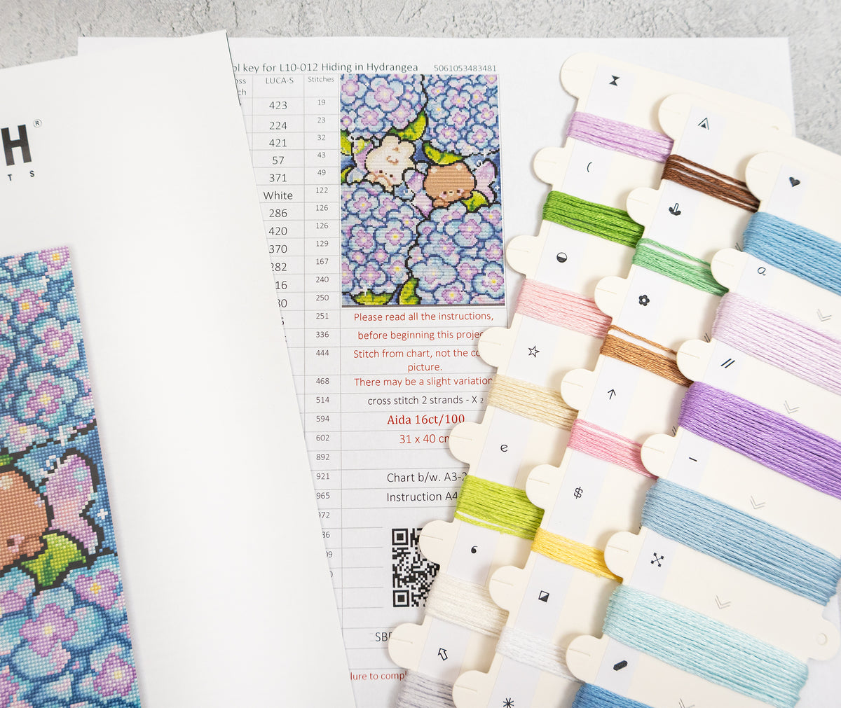 A cross-stitch kit laid out on a light gray surface, showing a printed pattern sheet with a floral design, a finished patch, multiple thread spools in assorted colors, and part of the instruction booklet.