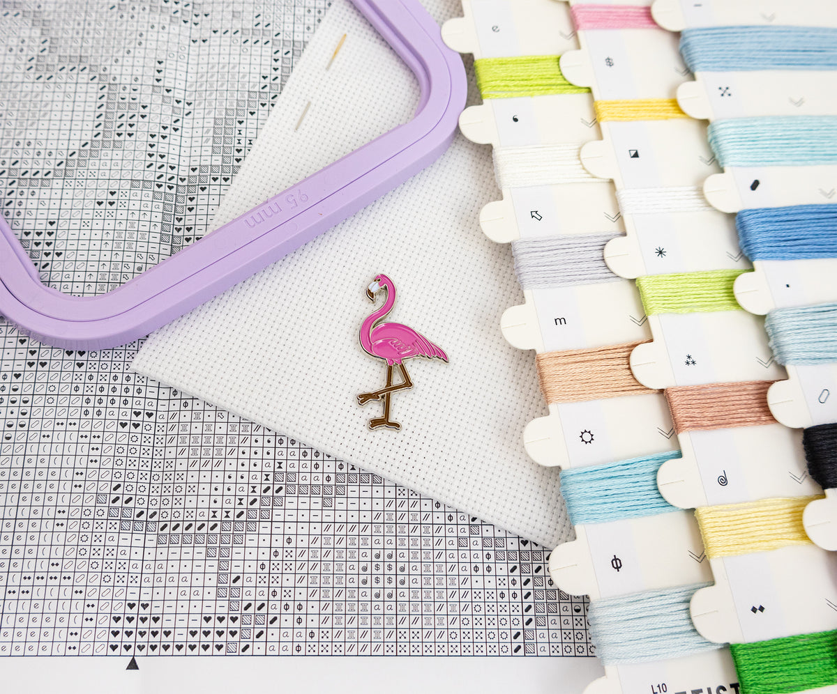 A pink flamingo-shaped enamel pin with a gold-toned leg stands on a white textured fabric next to a lavender embroidery hoop, a printed cross-stitch pattern sheet, and multiple spools of thread in assorted colors.