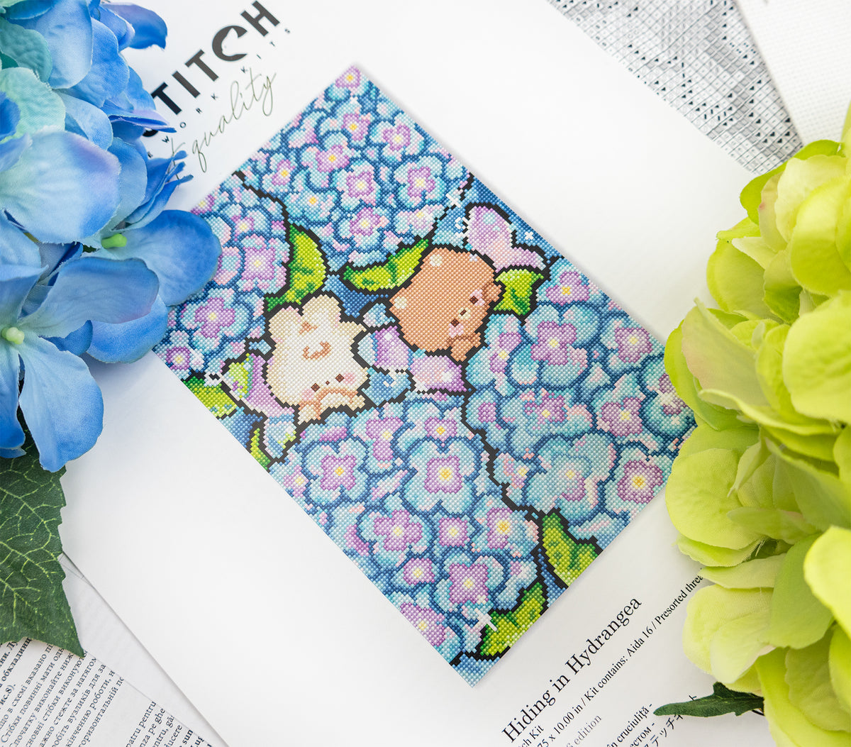 A square fabric featuring a cute pixel-art style design of two small animals among blue hydrangea flowers, styled on a white book page with blue and green hydrangea flowers around it.