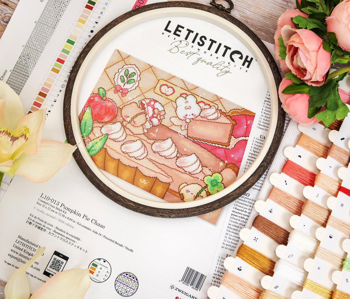 A cross-stitch design featuring a cozy autumn picnic scene with pastries, apples, and a basket, surrounded by thread spools, flowers, and printed craft materials on a wooden surface. The brand name LETISTITCH is visible at the top of the circular piece.