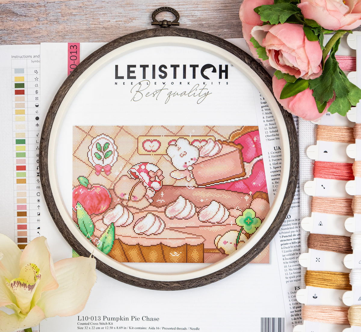 A framed cross-stitch design of a pumpkin pie with a cute pumpkin character, displayed in a wooden hoop on a styled flat-lay background with flowers, thread spools, and a color chart.