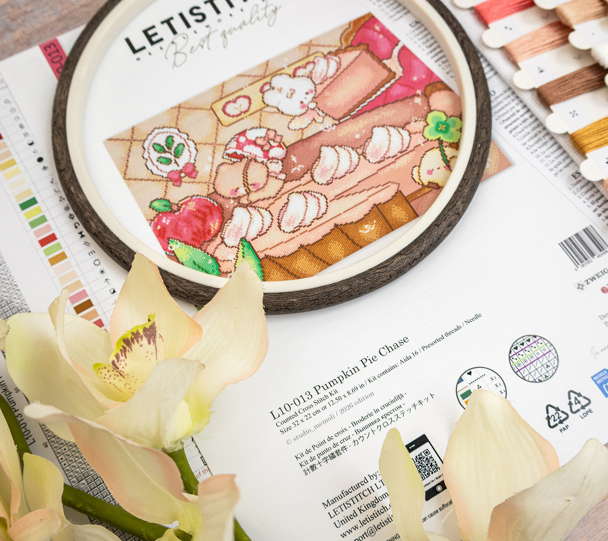 Embroidery kit containing a pumpkin pie design with cute cartoon characters, displayed on top of printed fabric or paper with thread spools and pale flowers in the foreground.