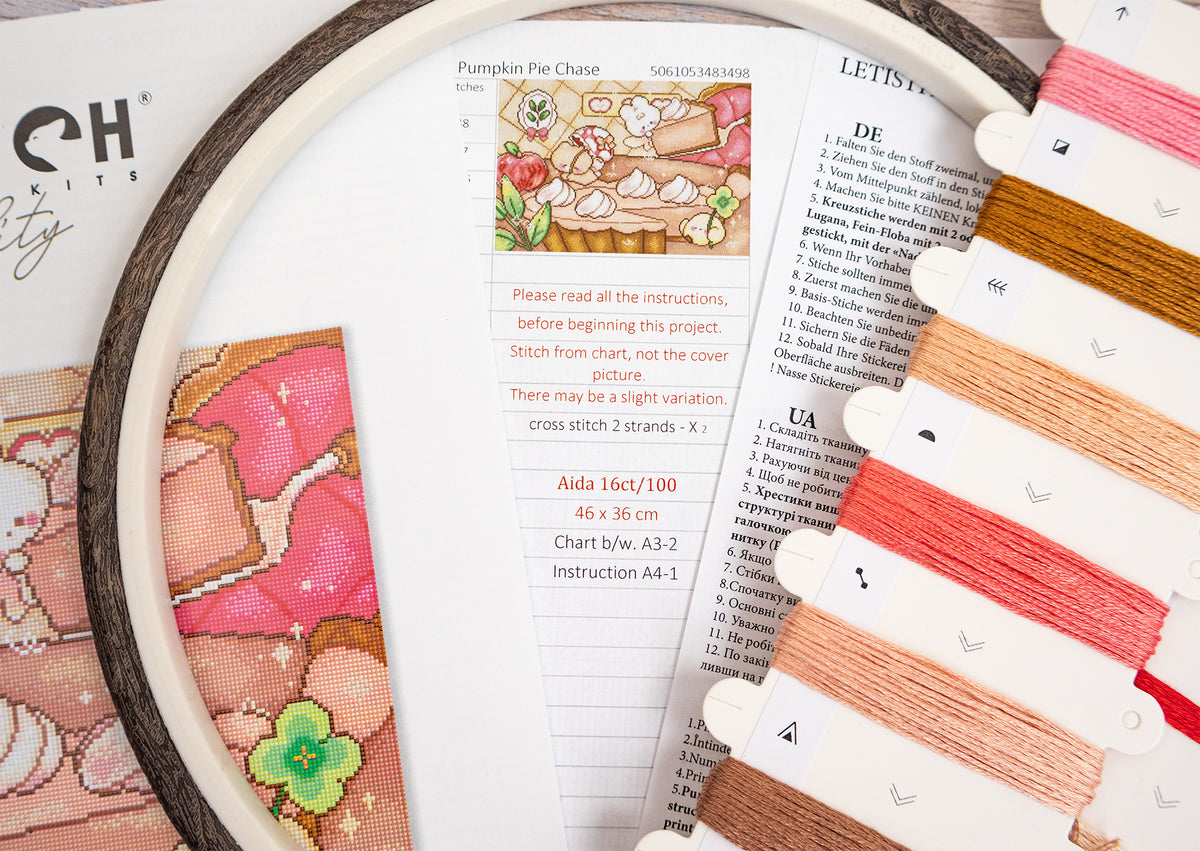 A close-up of a cross-stitch embroidery hoop containing a pumpkin pie design, with printed instructions and thread color swatches laid out beside it on a white surface.
