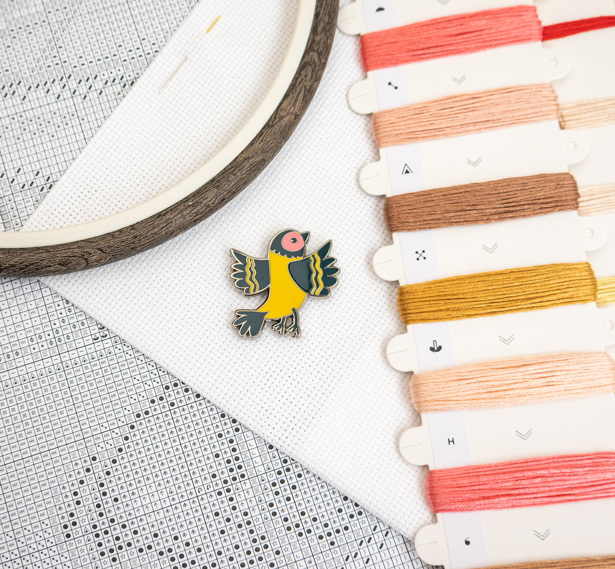 A small bird-shaped enamel pin in yellow, green, and pink is placed on white embroidery canvas next to an embroidery hoop and a row of colorful embroidery floss spools.