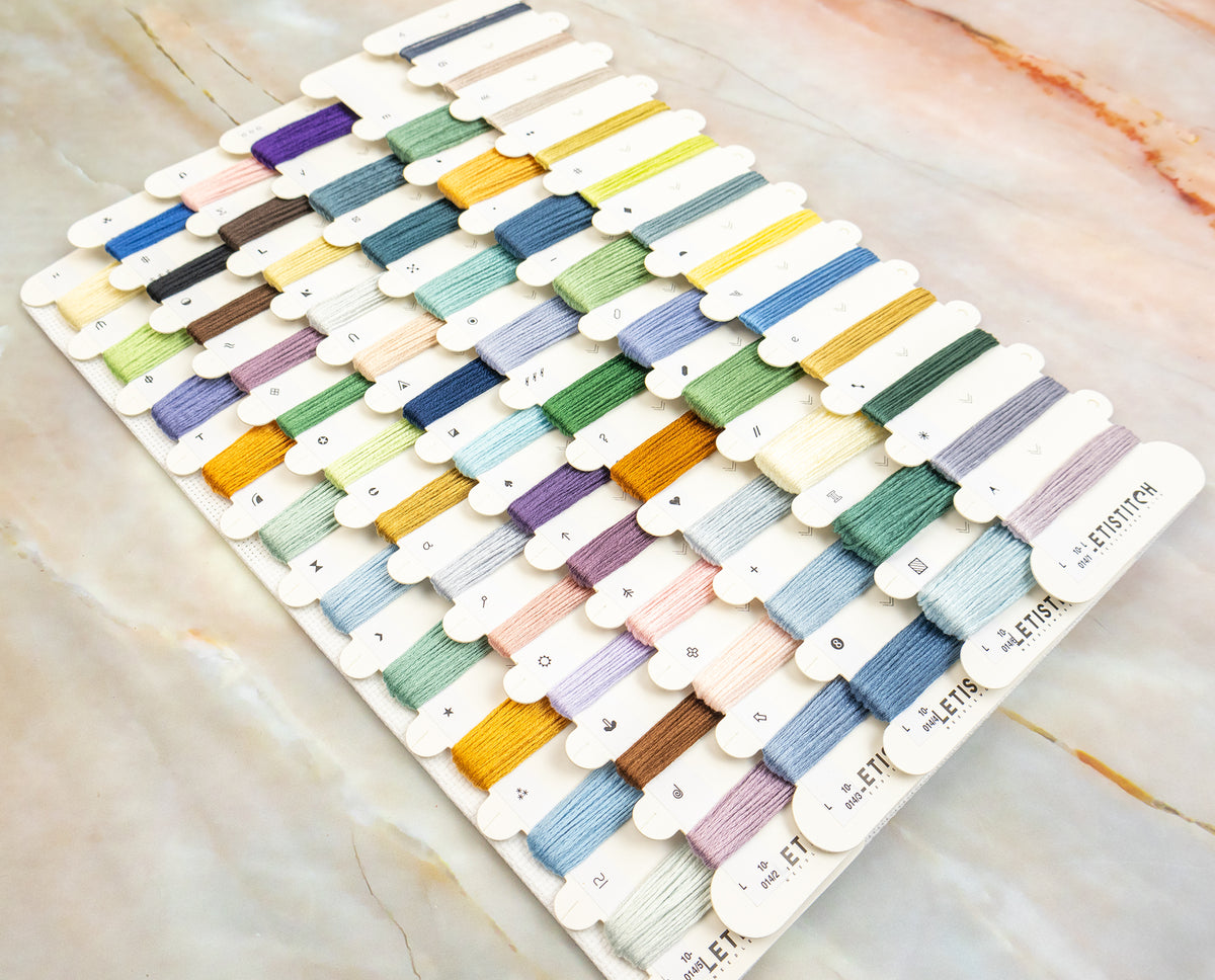 A large card displaying many small thread or yarn color samples arranged in rows on a marble-like surface. The card includes many pastel and muted tones plus some brighter colors