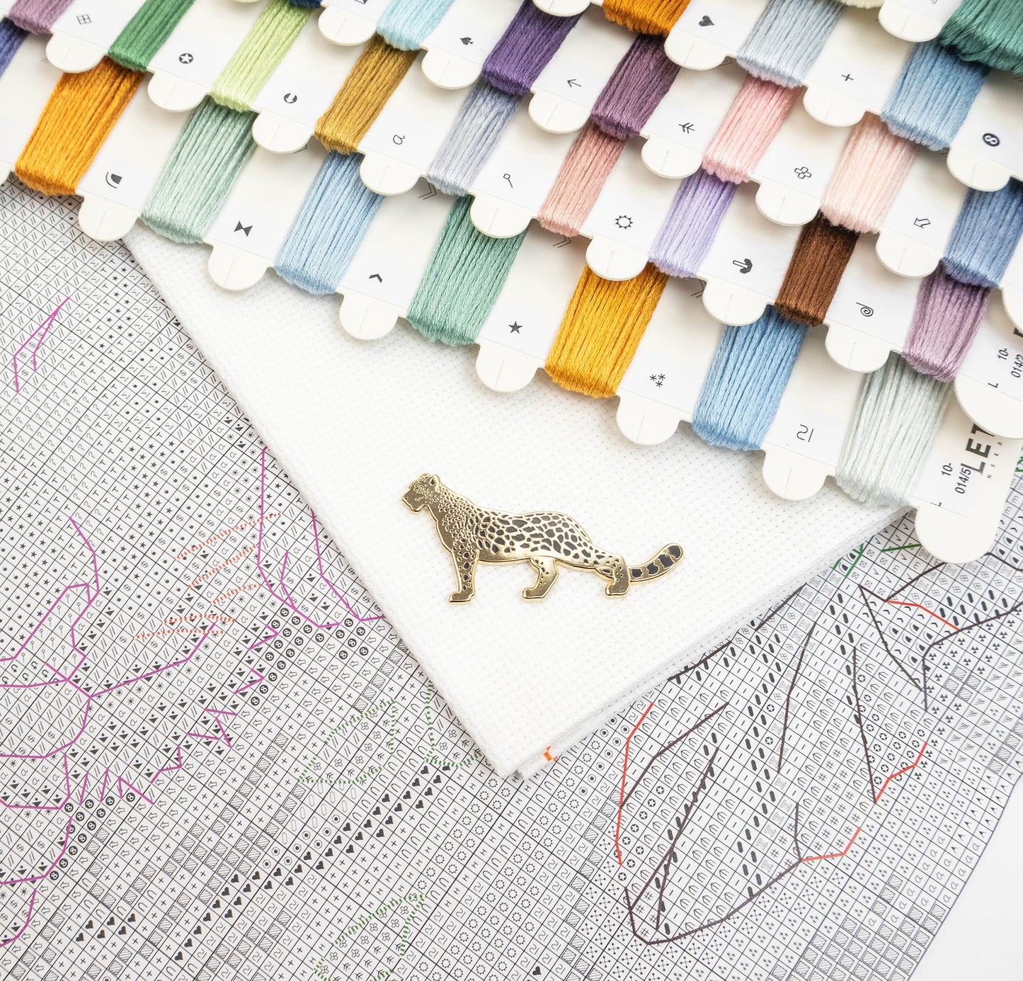 A close-up flat lay of a white embroidery canvas with a small leopard-shaped metal pin placed on it, surrounded by many thread spools in assorted colors and a counted square grid.