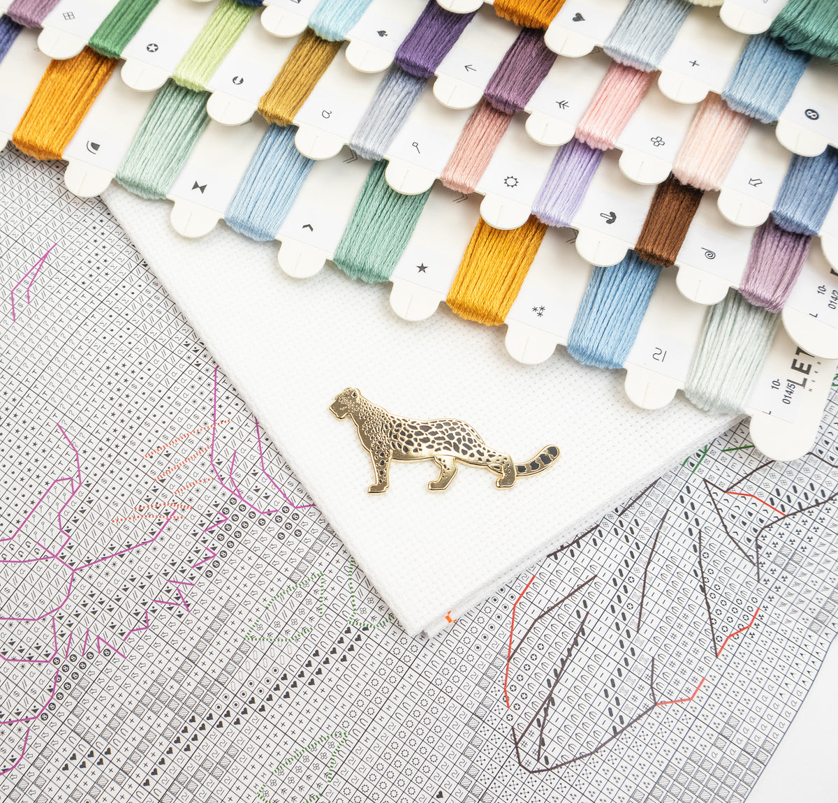A close-up flat lay of a white embroidery canvas with a small leopard-shaped metal pin placed on it, surrounded by many thread spools in assorted colors and a counted square grid.