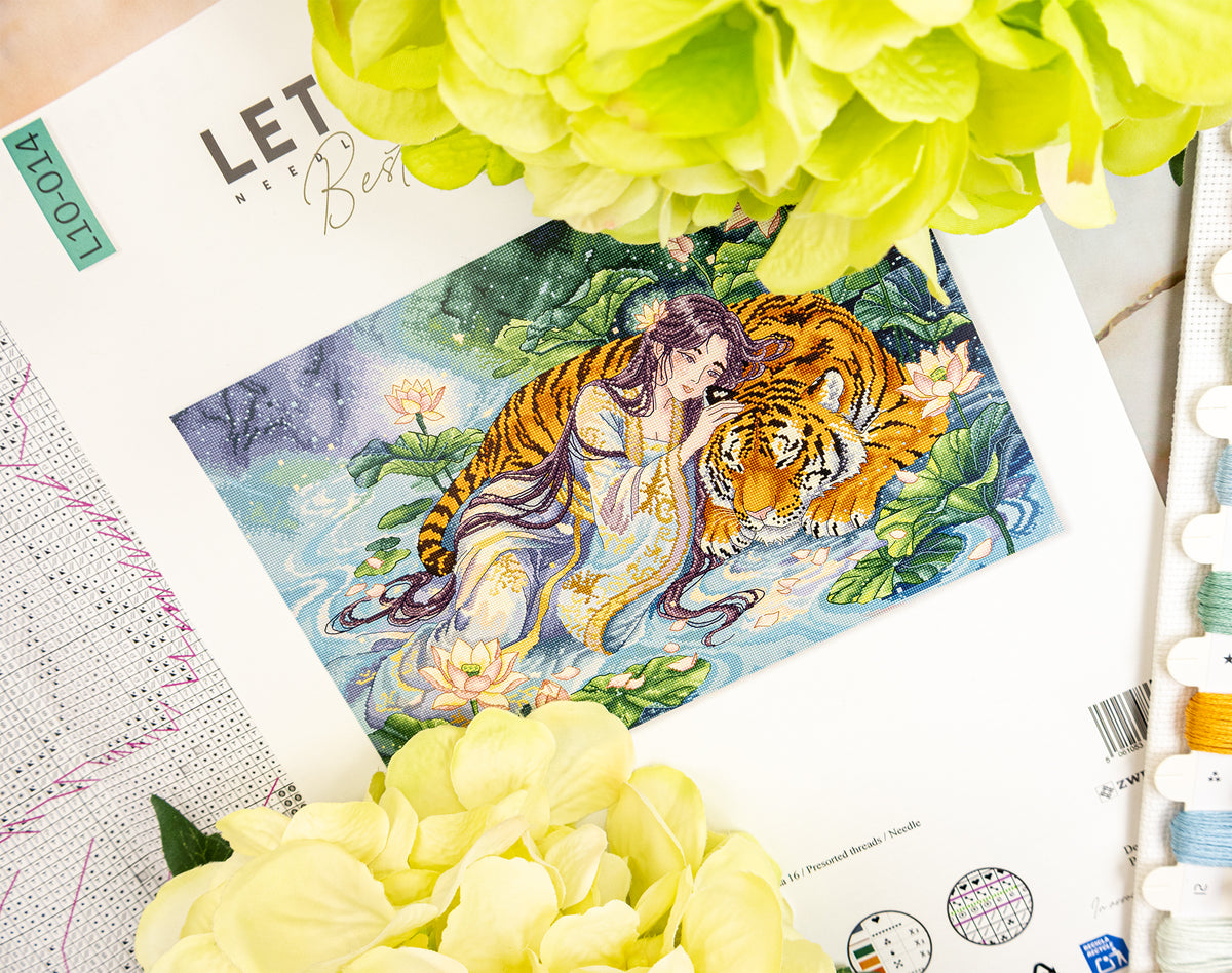 A printed cross stitch pattern sheet featuring an illustrated scene of a woman and a tiger among lotus flowers, styled with pale yellow flowers and thread spools on a light surface.