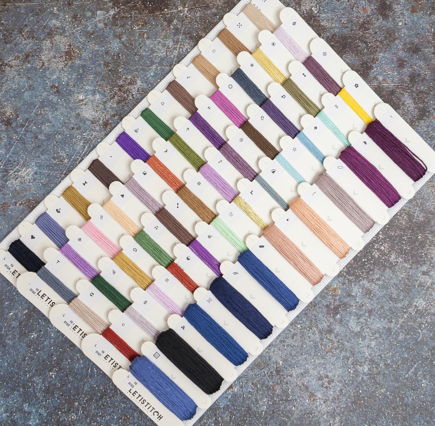 A board displaying many small thread or embroidery floss color samples, arranged in rows on a textured gray surface. The board includes many different colors rather than one dominant shade, so the color identity is best described as multicolor.