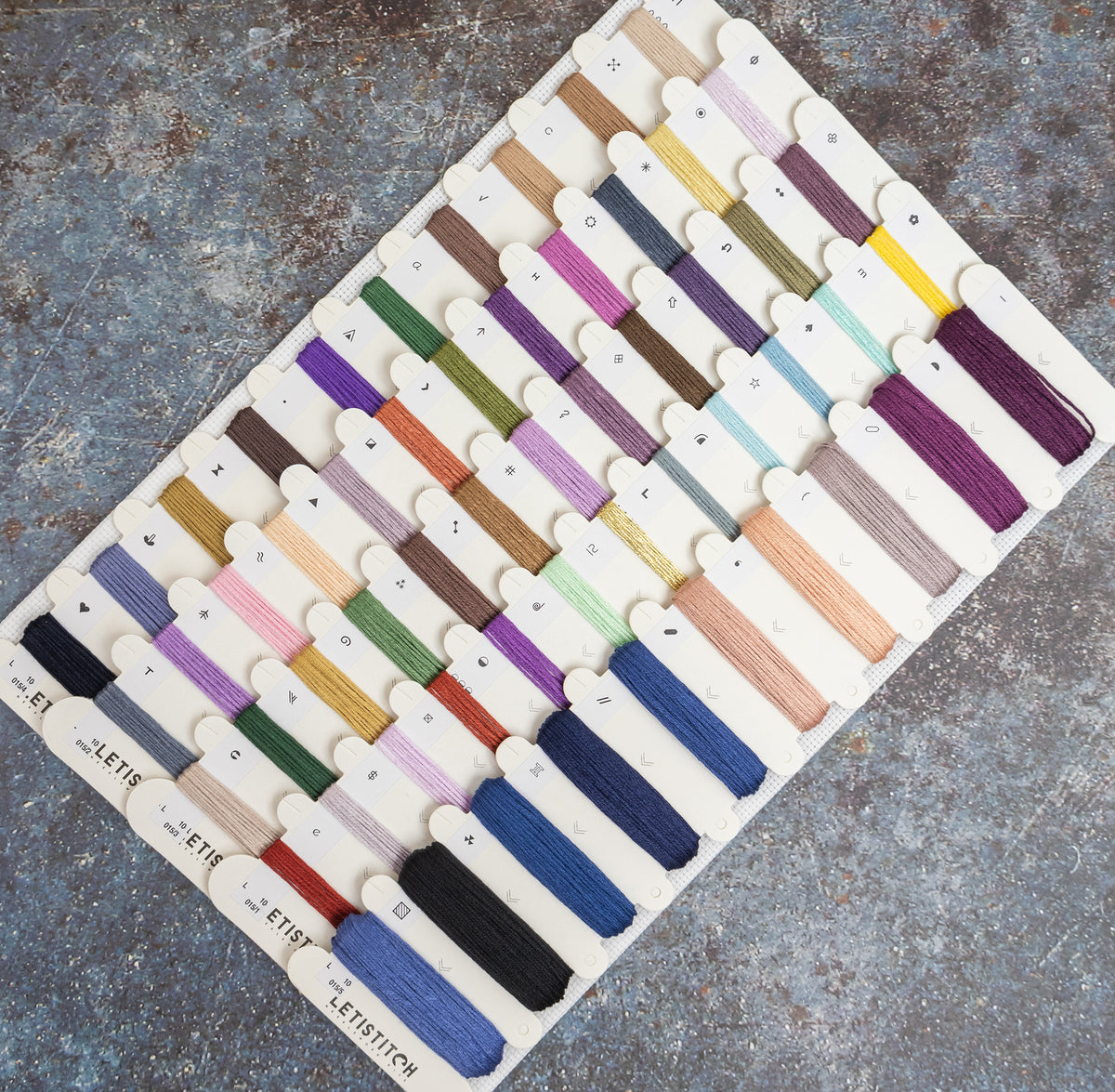 A board displaying many small thread or embroidery floss color samples, arranged in rows on a textured gray surface. The board includes many different colors rather than one dominant shade, so the color identity is best described as multicolor.