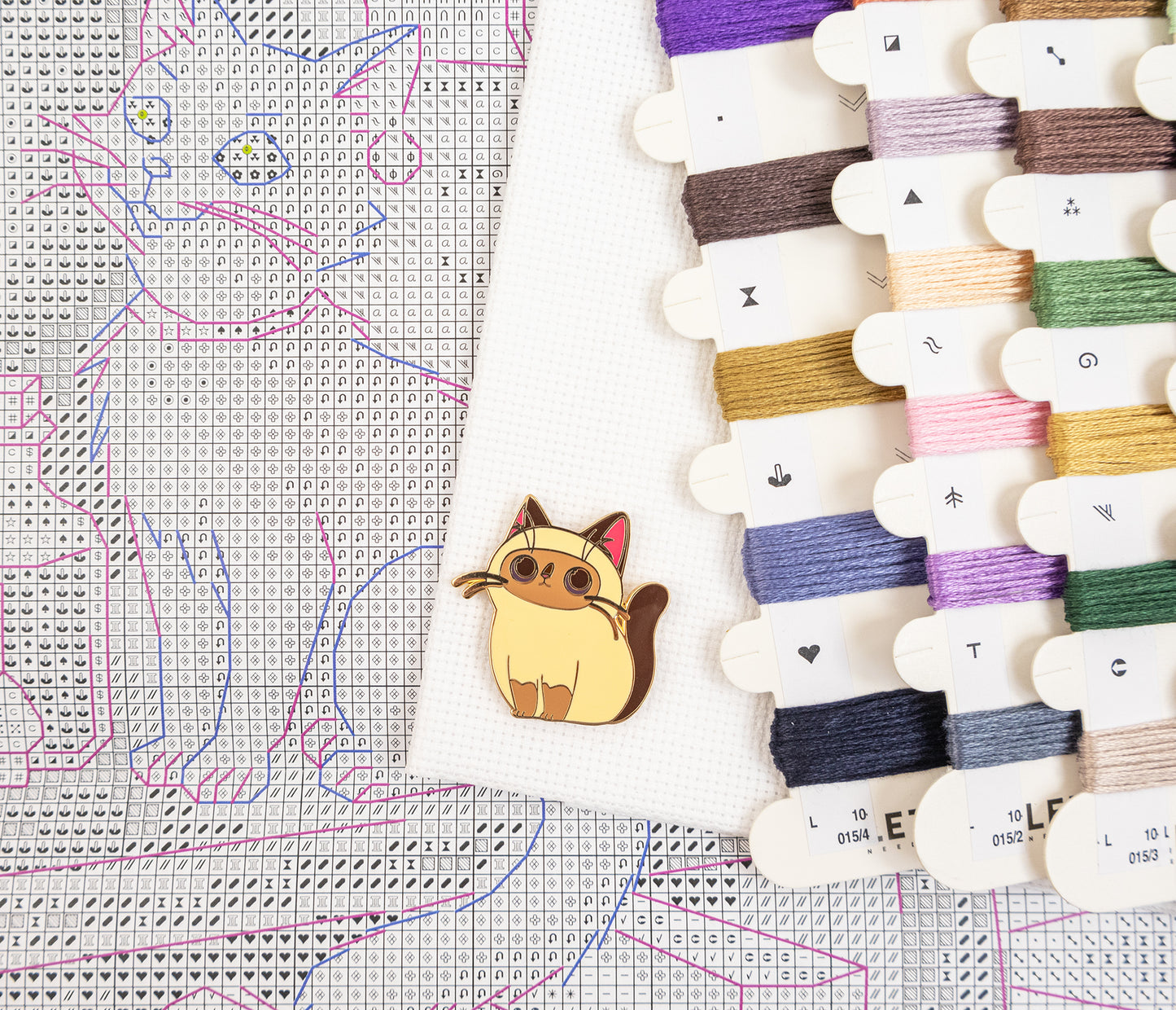 A small embroidered cat-shaped pin or charm in tan, cream, and pink lies on a white fabric sample. Surrounding it are multiple thread spools in assorted colors and a counted cross-stitch pattern sheet with a cat design.