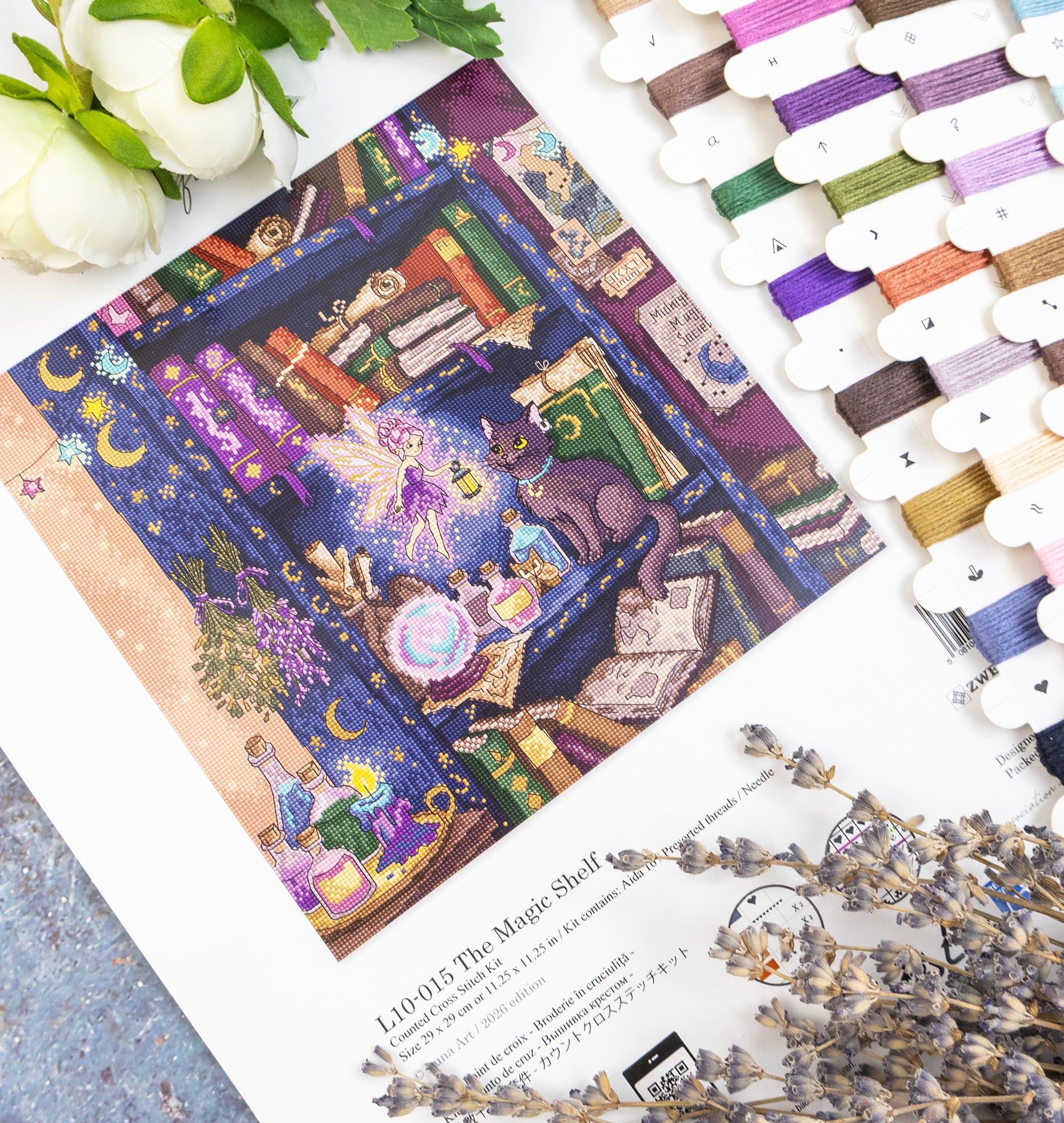A flat-lay product photo of a needlepoint canvas featuring a whimsical fantasy bookshelf scene with a fairy and a cat. The canvas is styled on a white surface with a book page underneath, surrounded by white roses, dried lavender, and a row of colorful thread spools on the right.