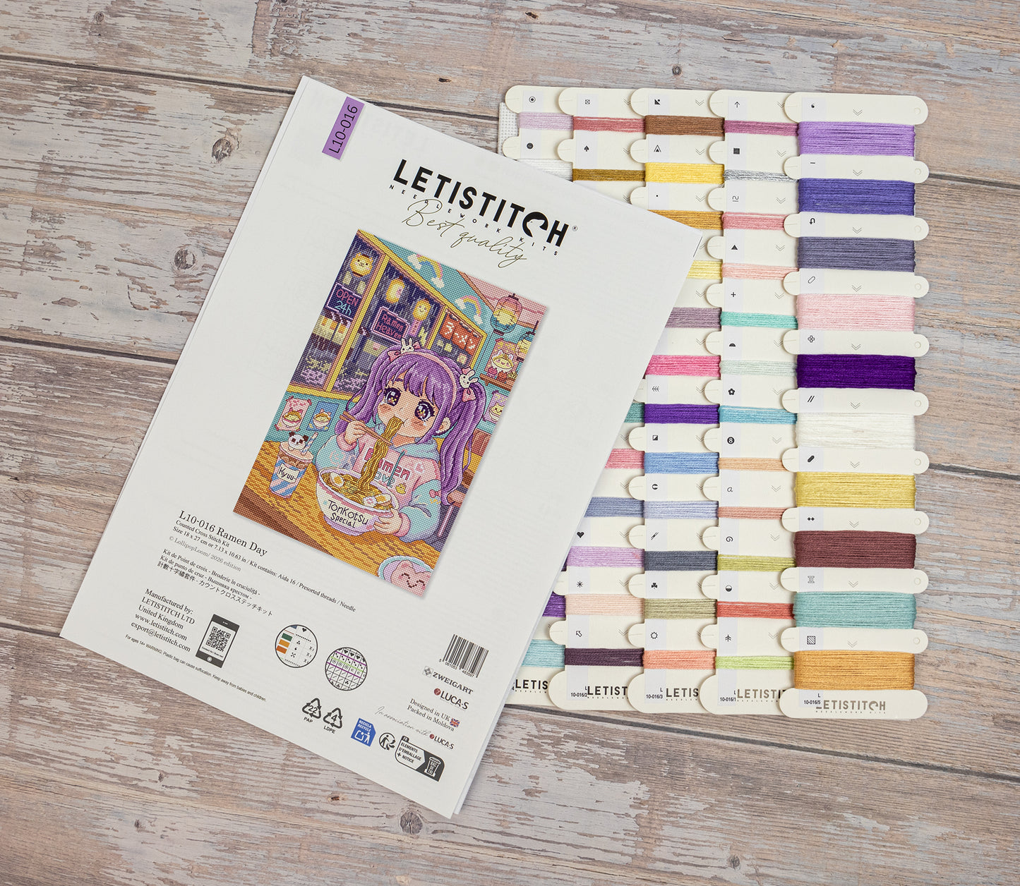 Letistitch Cross Stitch kit packaging and a matching thread sampler board displayed on a rustic wooden surface. The package features an illustration of a purple-haired girl in a cozy room, and the thread sampler shows multiple pastel and bright color options.