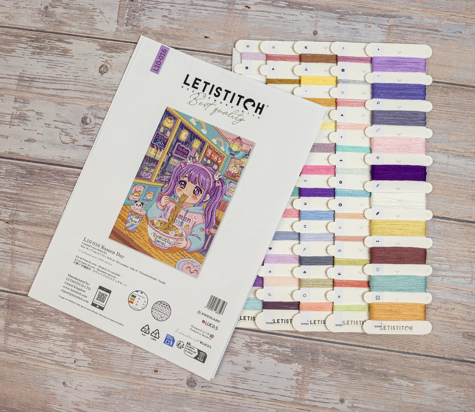 Letistitch Cross Stitch kit packaging and a matching thread sampler board displayed on a rustic wooden surface. The package features an illustration of a purple-haired girl in a cozy room, and the thread sampler shows multiple pastel and bright color options.