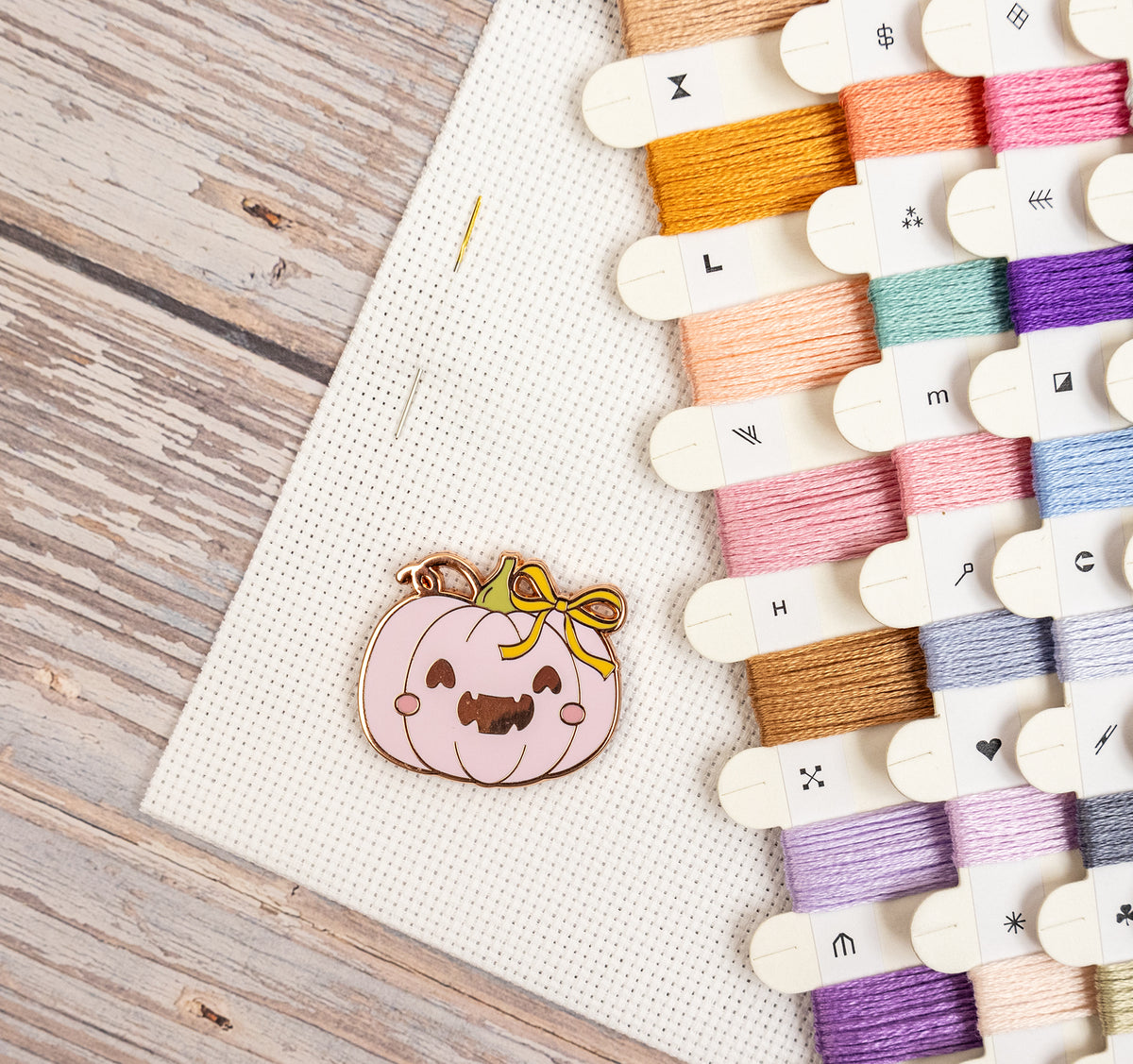 A cute pumpkin-shaped enamel pin with a smiling face and yellow bow, displayed on a white cross-stitch canvas next to a colorful spool of embroidery floss on a rustic wooden table.