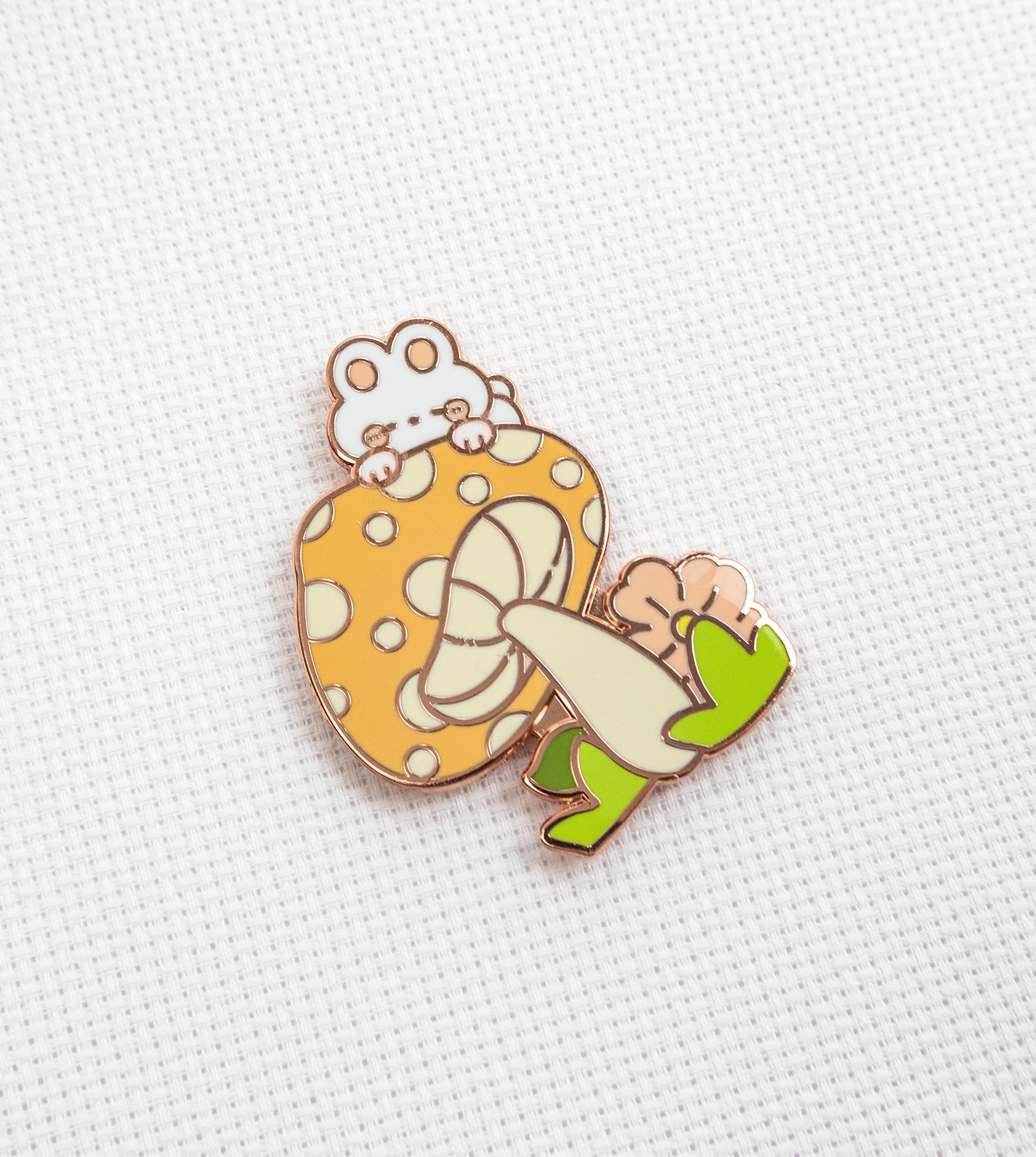 A cute enamel pin featuring a white cat peeking from behind a yellow strawberry with white spots, a cream-colored bow, and green leaves, displayed on a textured white fabric background.